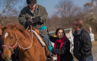 Colt Romberger watches father ride a horse while a woman assists him.