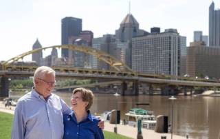 Couple stands outside and looks lovingly at each other in front large driving bridge. There is a river and a city in the background.