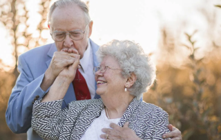 Walt and Margie Cook, a smiling senior couple in a field, Walt is kissing Margie's hand.