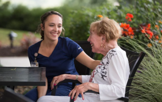 A nurse sits outside with a senior woman and they look at each other laughing.