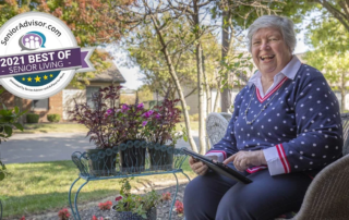 Smiling woman sitting on her porch with a tablet and smiling, Best of Senior living badge from Senior Advisor is in one corner.