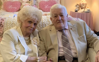 Evelyn and Carl Hackman, a senior couple seated together in front of a bed.