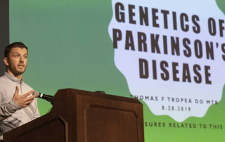 Dr. Tropea, a man speaking at a podium in front of slide labeled "Genetics of Parkinson's Disease."