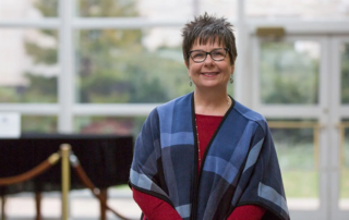 Carol Olnick, a smiling woman with short hair and glasses standing indoors.