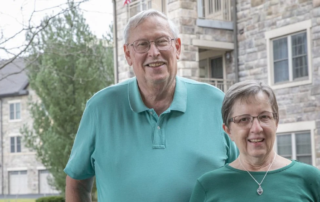 David and Janice Rohrbach, a smiling senior couple in front of a stone apartment building.