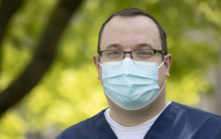 A man with short hair, glasses and a mask wearing blue scrubs.