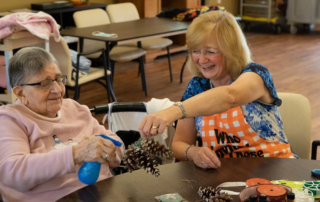 Smiling aide helping senior woman with pinecone crafts.