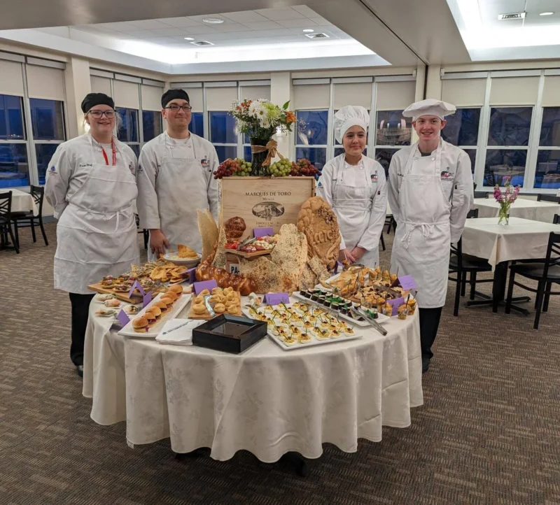 Group of smiling chefs behind table with delicious food and baked goods.