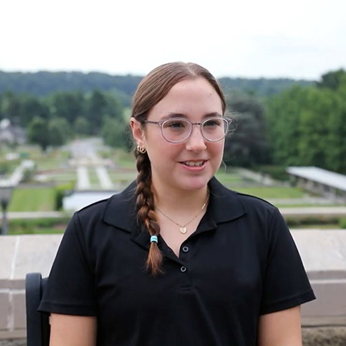 Smiling young woman with braided hair abd glasses, in a black shirt, standing outdoors.