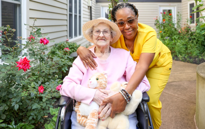 Smiling senior woman in a wheelchair being hugged by a smiling nurse in bright scrubs outdoors in a garden.