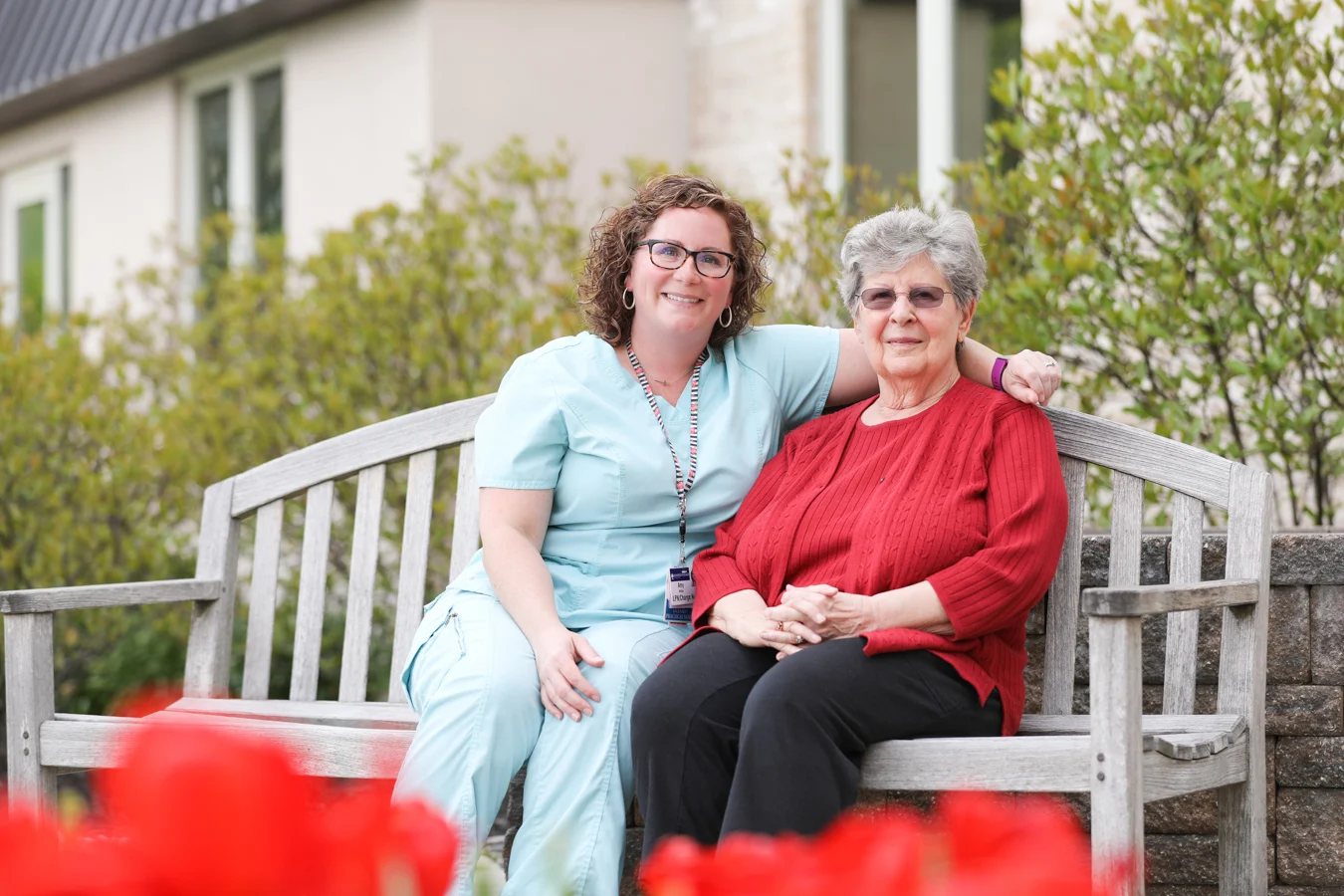 Senior woman and nurse seated on a wooden bench outdoors together.