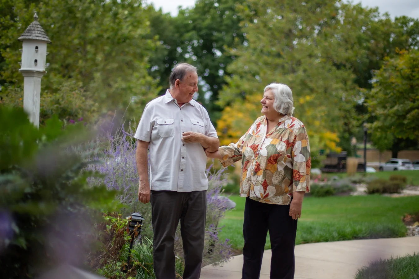 IMG_7223 Smiling senior couple linking arms while walking through gardens outdoors.