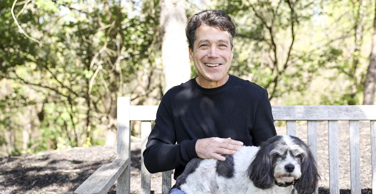 Ron-Piroli Smiling mature man seated on a wooden bench outdoors with a dog in his lap.