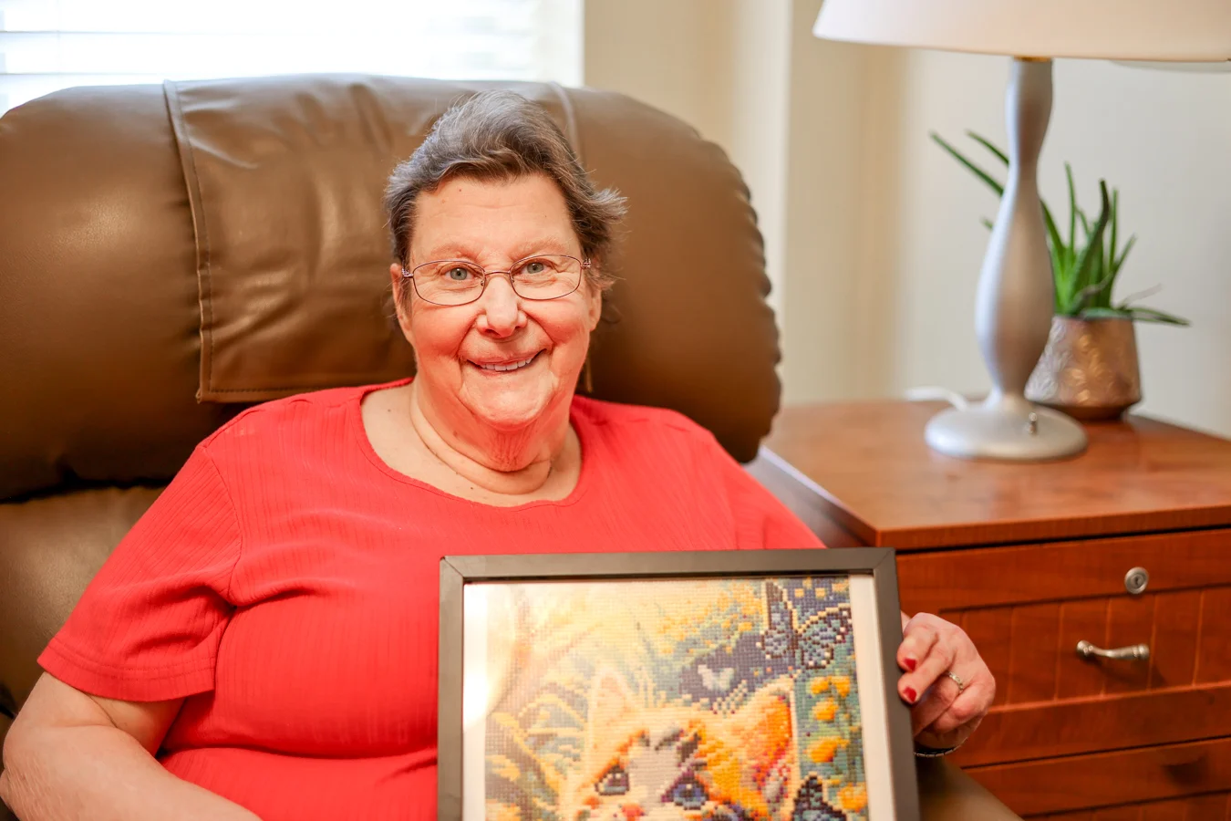 Smiling senior woman holding up a piece of art while seated in a plush chair.