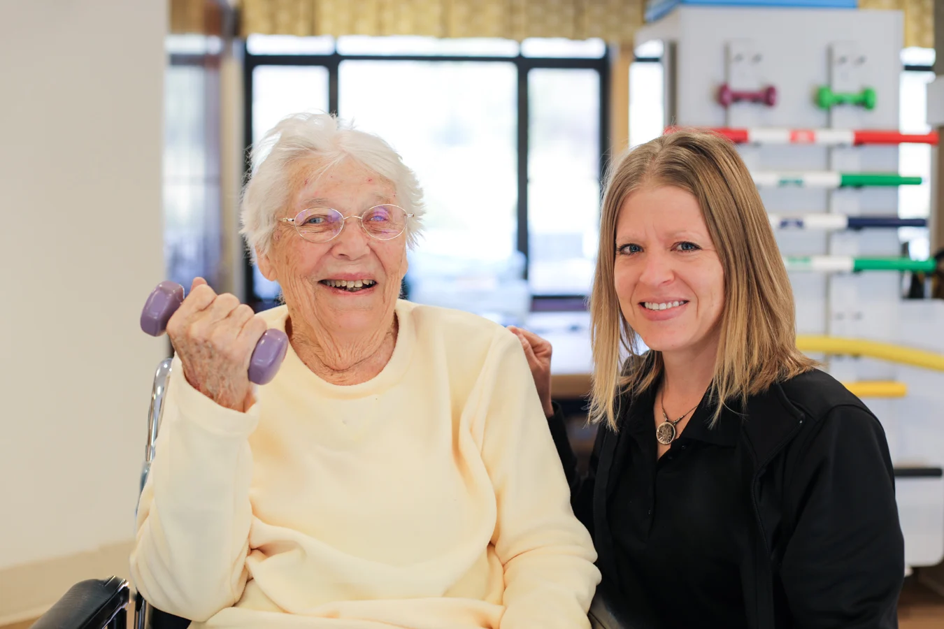 Smiling senior woman lifting weight while seated beside a physical therapist.