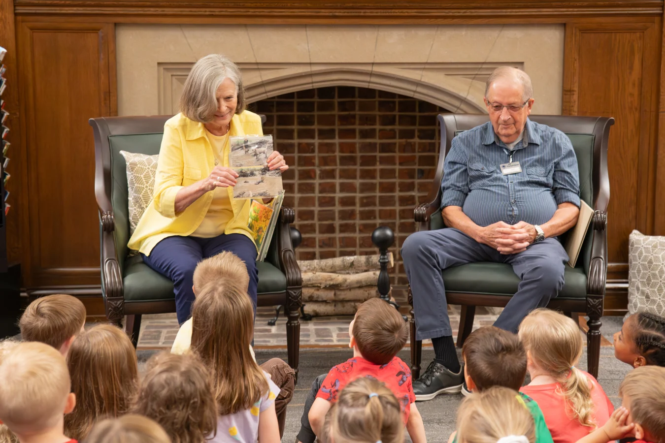 Senior volunteers seated in chairs showing off pictures to a large group of children.