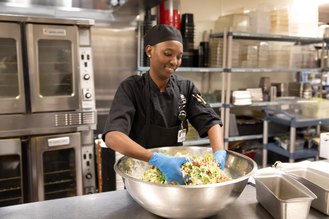 Smiling woman in chef's attire preparing a salad in a large bowl in a kitchen.