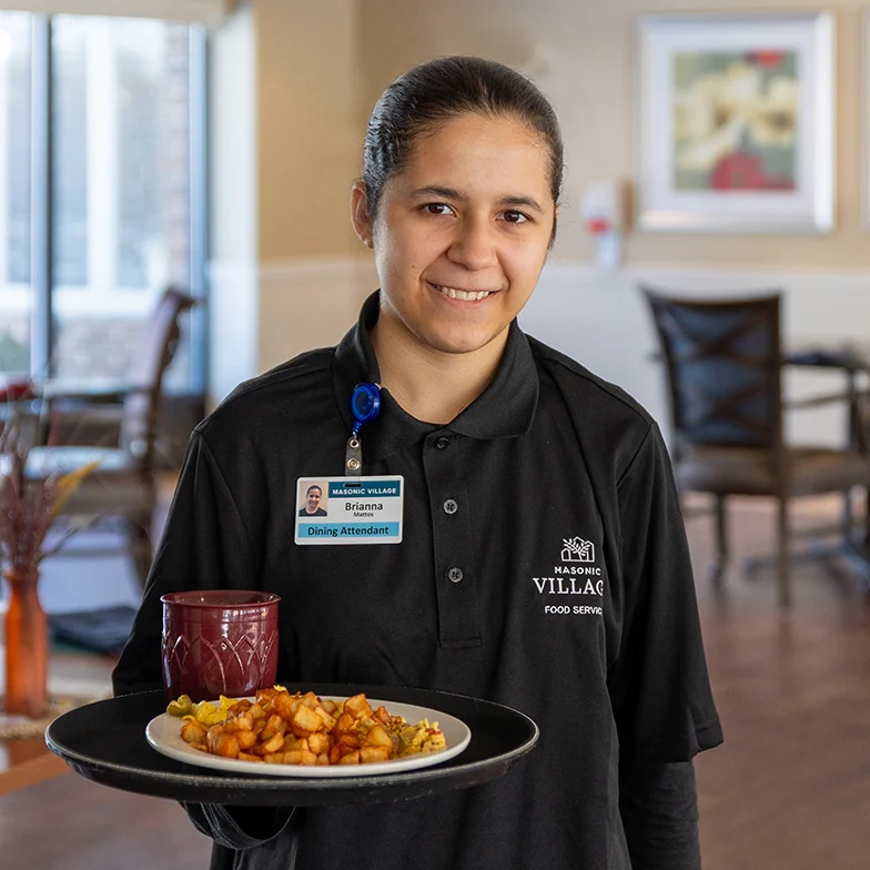 Smiling young woman in a black Masonic Village shirt, holding a plate of food while standing in a dining room.