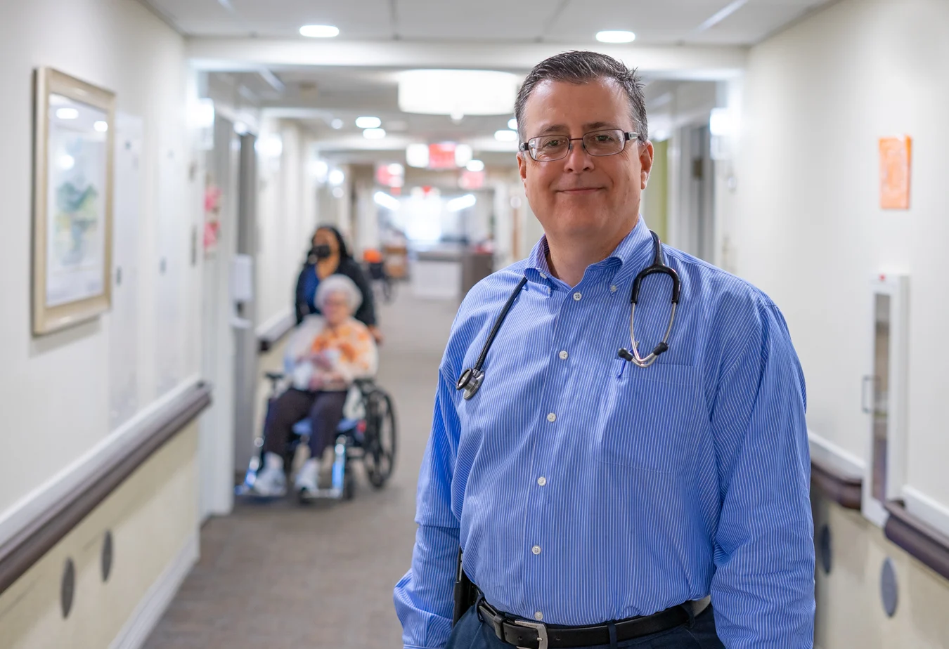 Smiling doctor with a stethoscope around his neck, standing in a well-lit hallway.