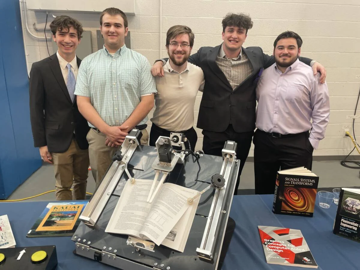 Group of young men standing together indoors. a table in front of them has books and machinery displayed.