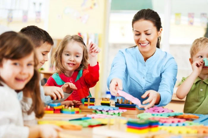 Smiling teacher helping group of toddlers with a puzzle at a table.