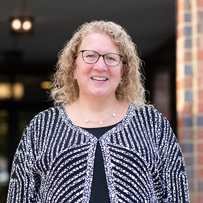 IMG_2357 Smiling woman with curly blond hair and glasses standing outdoors.