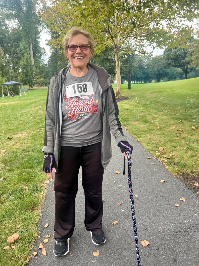 Smiling woman with a runners bib walking on a path with a cane.