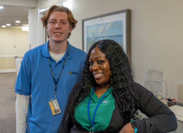 Smiling nurse and student standing together at a station in a hallway.