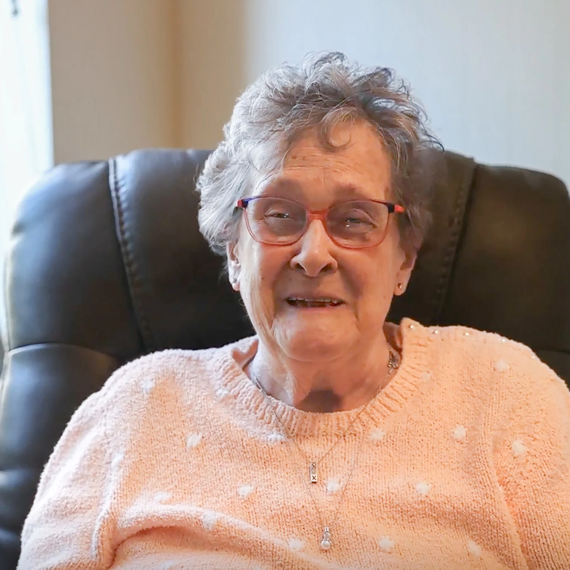 Smiling senior woman with glasses seated in a brown chair.